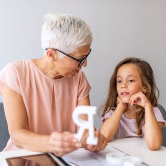 Eine Frau mit weißen Haaren zeigt einem Mädchen in der Sprachtherapie ein großes R.