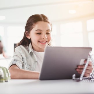 Ein junges Mädchen sitzt fröhlich vor einem Laptop im Klassenzimmer.