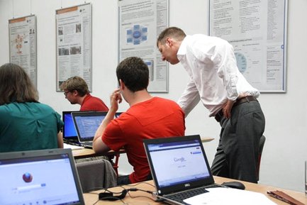 [Translate to English:] People are sitting in a classroom. One person is standing behind another and explaining something to the other person.