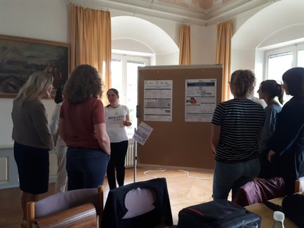 Mehrere Personen stehen in einem Raum mit hohen Fenstern. Eine Person zeigt auf eine Pinnwand mit mehreren angehefteten Blättern, die anderen Personen stehen in einem Kreis um die Pinnwand und hören zu.
