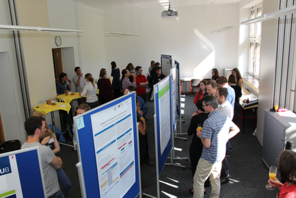 Mehrere Personen stehen in einem hellen Raum mit großen Fenstern. Im Raum sind mehrere blaue Stellwände mit Informationspostern aufgestellt. Die Personen sind in kleinen Gruppen verteilt und unterhalten sich.